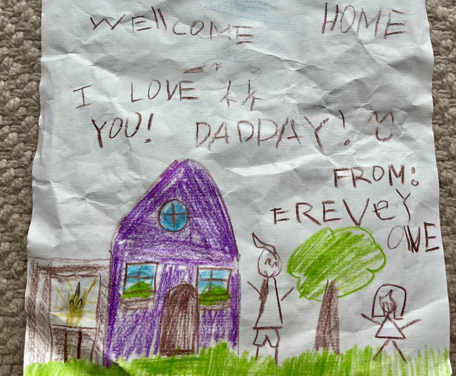 A welcome-home drawing of a purple house, a tree, and figures, with the message “Welcome home! I love you! Dadday!” from the kids.